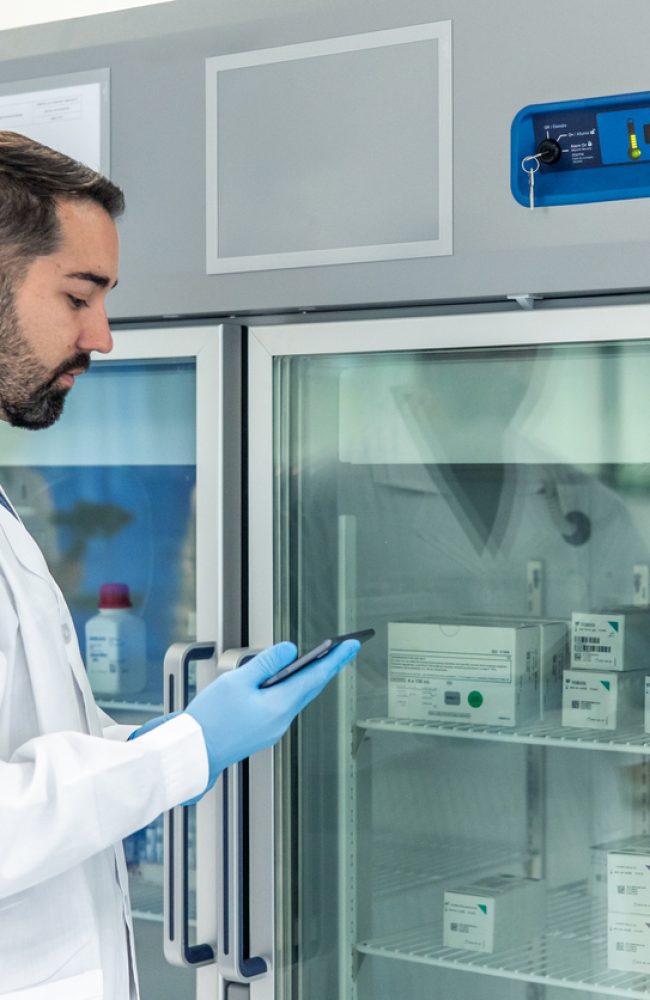 Scientist wearing a lab coat and gloves is using a digital tablet to check inventory or record data related to medical samples stored in a specialized laboratory refrigerator